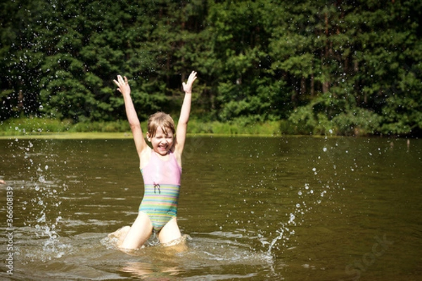 Fototapeta a little girl in a bathing suit jumps and splashes water in the lake in summer