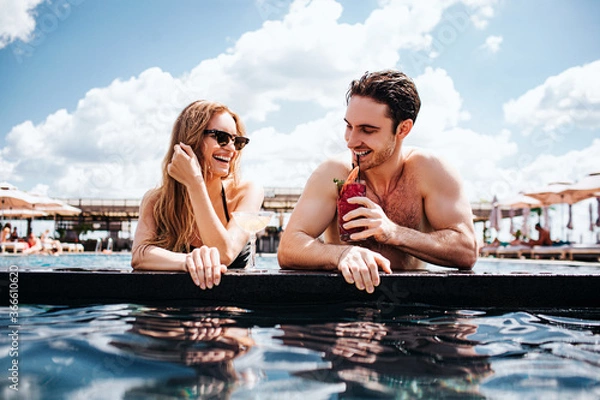 Fototapeta Young hot couple resting at swimpool. Low view of man and woman enjoy rest in resort spa hotel. Look at each other and smile. Drinking cocktails. Summertime vacation.