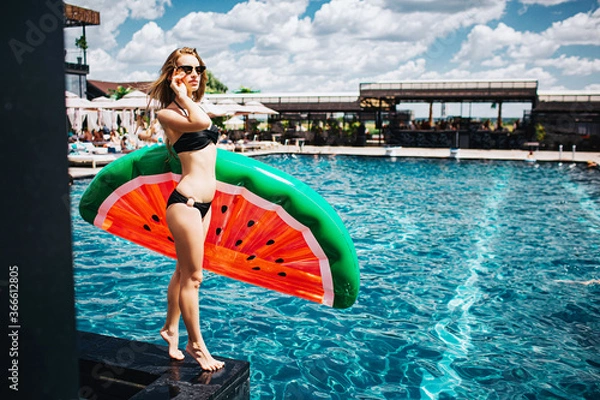 Fototapeta Young hot woman resting at swimpool. Slim well-built girl posing with swimming ring in water melon shape in hands and touch sunglasses. Rest relax in resort.