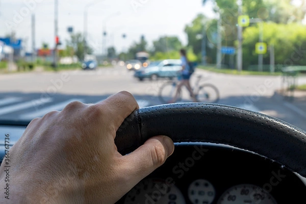 Fototapeta the driver hand at the wheel of a car against the background of a pedestrian crossing and a pedestrian girl bicyclist crossing the road