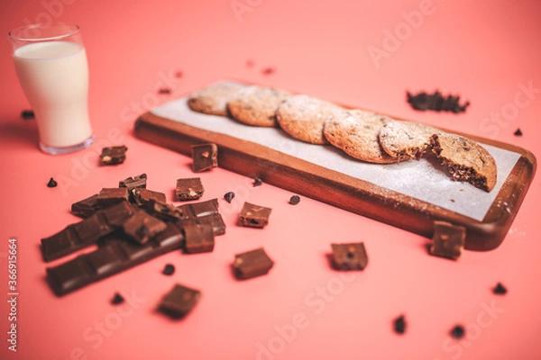 Fototapeta Freshly made Chocolate Chip Cookies on a wooden serving board. Cookies with a glass of fresh milk on the side along with chocolate chips and pieces of chocolate around on a pink background.Freshly mad