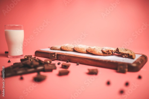 Fototapeta Freshly made Chocolate Chip Cookies on a wooden serving board. Cookies with a glass of fresh milk on the side along with chocolate chips and pieces of chocolate around on a pink background.Freshly mad