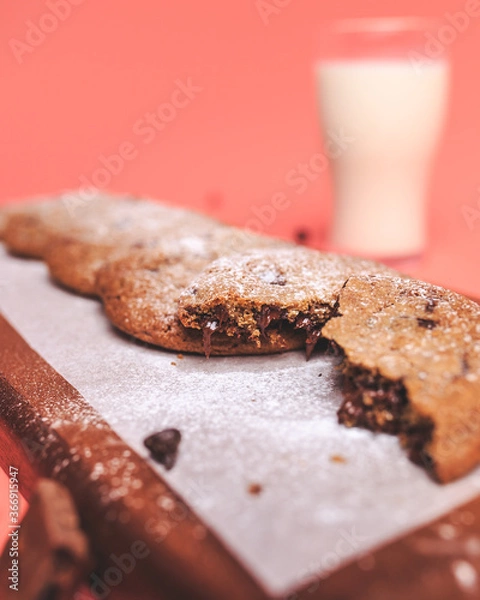 Fototapeta Freshly made Chocolate Chip Cookies on a wooden serving board. Cookies with a glass of fresh milk on the side along with chocolate chips and pieces of chocolate around on a pink background.Freshly mad