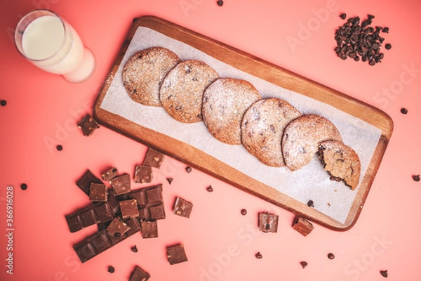 Fototapeta Freshly made Chocolate Chip Cookies on a wooden serving board. Cookies with a glass of fresh milk on the side along with chocolate chips and pieces of chocolate around on a pink background.Freshly mad