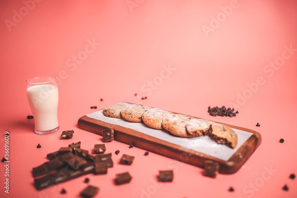 Fototapeta Freshly made Chocolate Chip Cookies on a wooden serving board. Cookies with a glass of fresh milk on the side along with chocolate chips and pieces of chocolate around on a pink background.Freshly mad