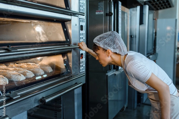 Obraz Young caucasian woman baker is looking at the bread baker process in an electric oven at a baking manufacturing factory.