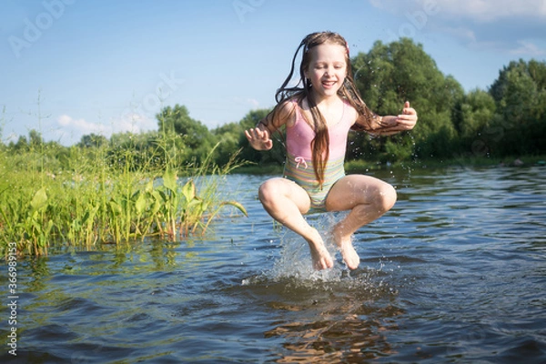 Fototapeta a little girl in a bathing suit jumps and splashes water in the lake in summer