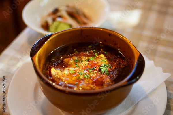 Obraz 陶器の茶色い器に盛られた、オニオングラタンスープ。日の光。背景にサラダ。レストランのテーブル。太陽光。