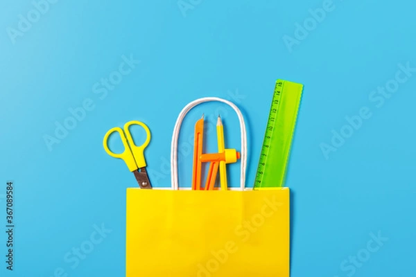 Fototapeta school supplies in a bright yellow paper bag on a blue background. Back to school concept. Concept of shopping in a stationery store. School sale concept. Top view, flat lay.