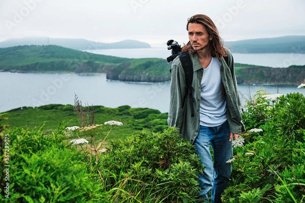 Fototapeta close up young long-haired man with a backpack and a photo tripod walking along a trail through the grass at the edge of a cliff with sky and sea background.. Travel and outdoor concept.