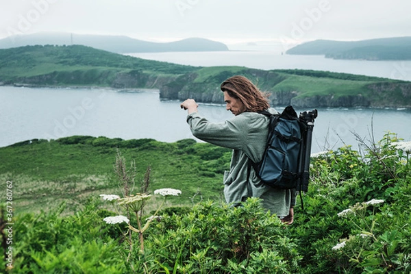 Fototapeta close up young long-haired man with a backpack and a photo tripod walking along a trail through the grass at the edge of a cliff with sky and sea background.. Travel and outdoor concept.