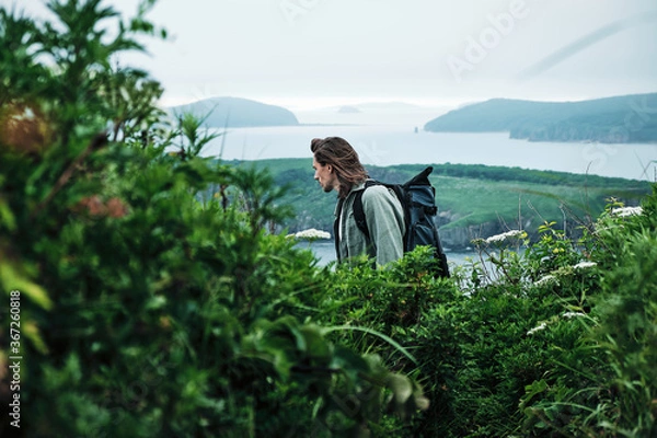 Obraz close up young long-haired man with a backpack and a photo tripod walking along a trail through the grass at the edge of a cliff with sky and sea background.. Travel and outdoor concept.
