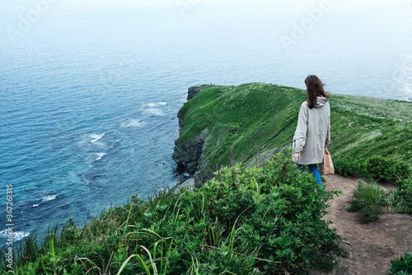 Obraz back view of a girl walking along a trail through the grass at the edge of a cliff with sky and sea background. Travel and vacation concept.
