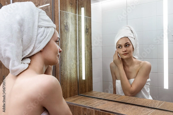 Fototapeta Young woman after a shower standing in the bathroom in front of the mirror with a towel wrapped on her head and washes her face with a cosmetic cotton pad with lotion. Skin care