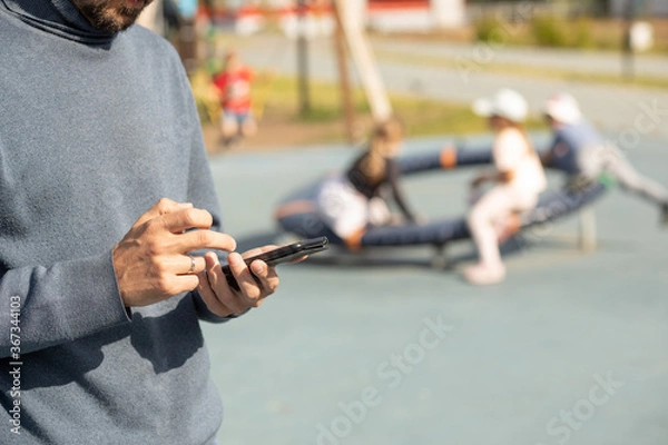 Fototapeta Dad looks at his phone and does not follow children in the playground