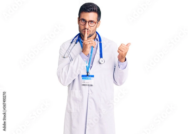Fototapeta Young hispanic man wearing doctor uniform and stethoscope asking to be quiet with finger on lips pointing with hand to the side. silence and secret concept.