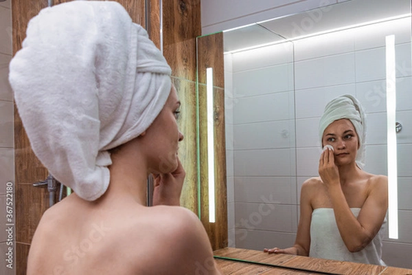 Obraz Young woman after a shower standing in the bathroom in front of the mirror with a towel wrapped on her head and washes her face with a cosmetic cotton pad with lotion. Skin care