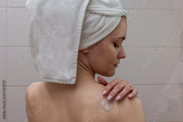 Obraz Beautiful young woman with a white towel on her head standing in the bathroom and applying shower foam to her shoulder.