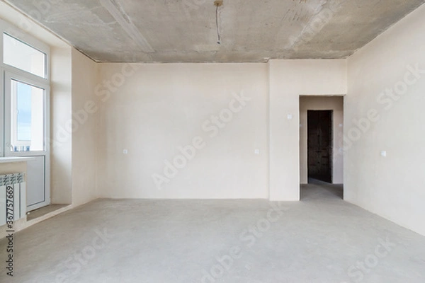 Fototapeta The front view an unfinished residential apartment with the white plastered walls, the empty doorway and the plastic balcony door