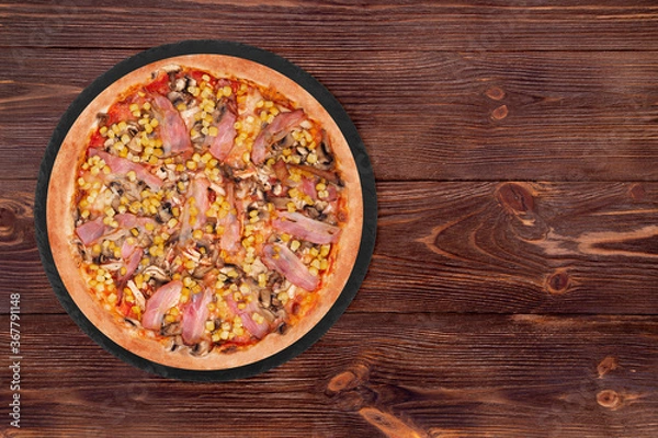 Fototapeta Delicious pizza with chicken breast, corn, bacon and mushrooms, on a round slate plate which is on wooden table, top view and copy space