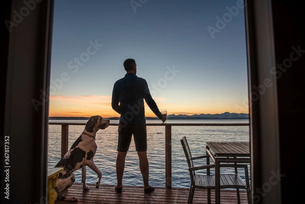 Fototapeta Man with dog framed by window enjoying sunset on seaside deck while holding glass of wine. 