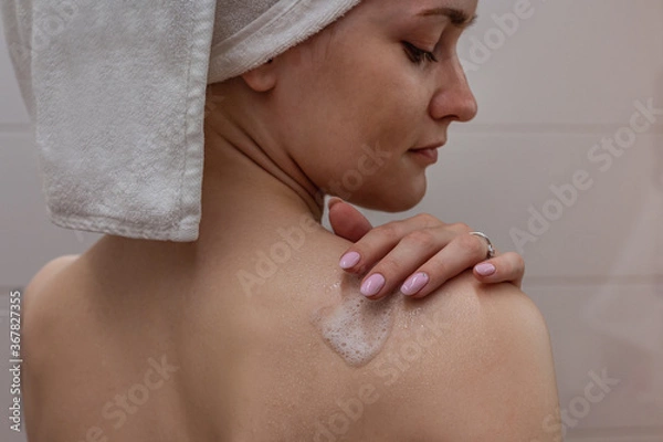 Obraz Beautiful young woman with a white towel on her head standing in the bathroom and applying shower foam to her shoulder.