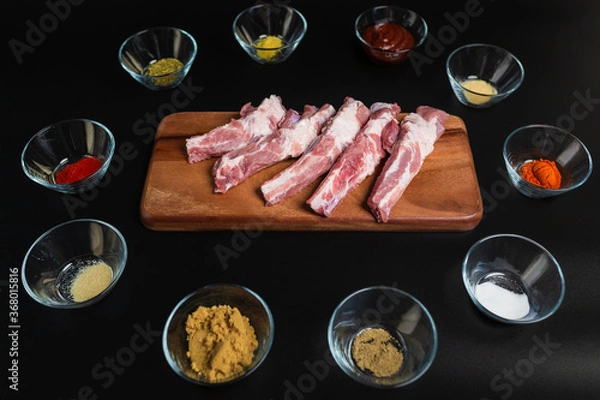 Fototapeta Close-up Raw Pork Ribs on a wooden board with lots of marinating spices around on a black background with free space for your text/decoration.