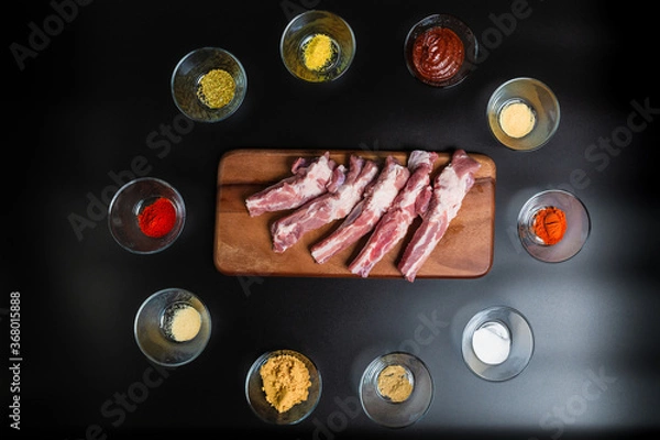 Fototapeta Close-up Raw Pork Ribs on a wooden board with lots of marinating spices around on a black background with free space for your text/decoration.