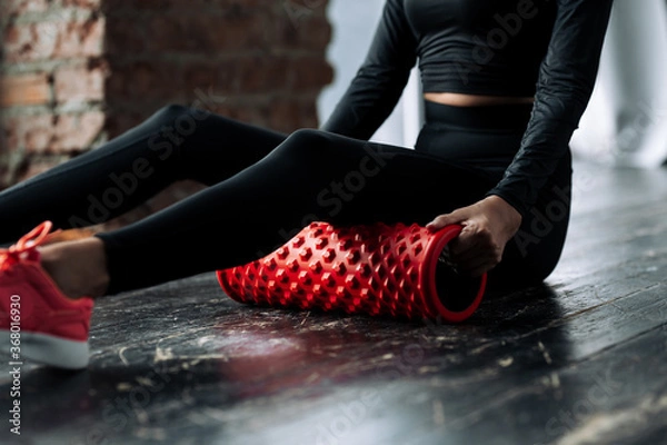 Fototapeta Close-up of a young woman in a black tracksuit performs a warm-up exercise by putting a roller massager for the fascia of the muscles of the body under the hips.