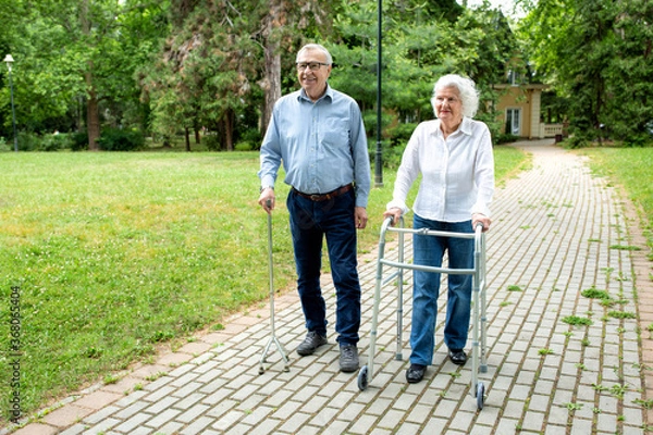 Fototapeta Senior man using a walking cane accompanied by a senior lady strolling with folding walker