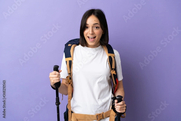Fototapeta Young caucasian woman with backpack and trekking poles isolated on blue background with surprise facial expression