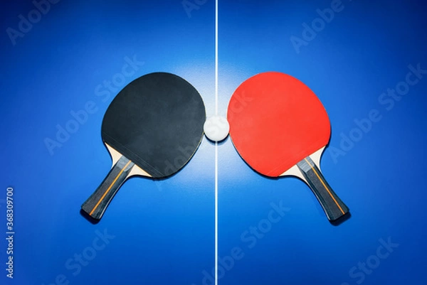 Fototapeta Top view black and red table tennis racket and a white ping pong ball on the blue ping pong table with a bright spotlight, Two table tennis paddle is a sports competition equipment for indoor exercise