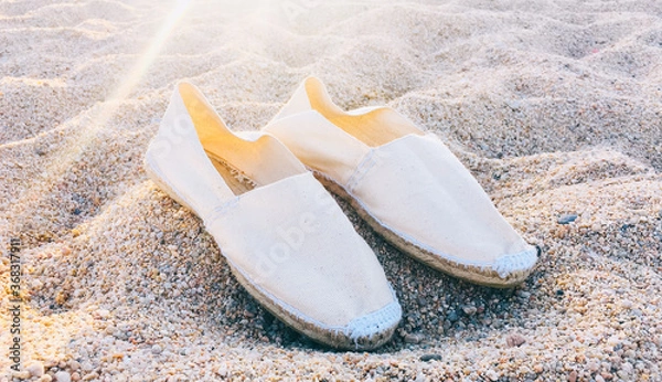 Fototapeta Nice white summer shoes on the sand of the beach with the rays of the sun. Espadrilles handmade in Spain with fabric.