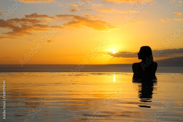 Obraz the Girl is standing back in the pool, slightly turning her head, her silhouette is reflected in the water. Against the background of the sunset and the ocean.