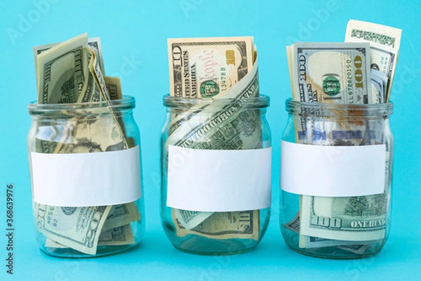 Fototapeta Three Piggy Bank from a Glass Jar for Life. New dollars sticking out of piggy banks on a blue background. The banks have white sheets for inscriptions. Mockup. Copy space. Economy concept