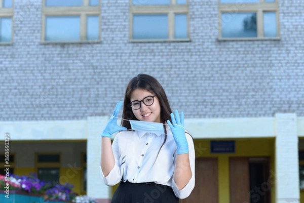 Obraz A schoolgirl wearing medical gloves and wearing a mask stands in front of the school. A high school student wearing protective gloves holds a medical mask. The girl in the defense smiles.