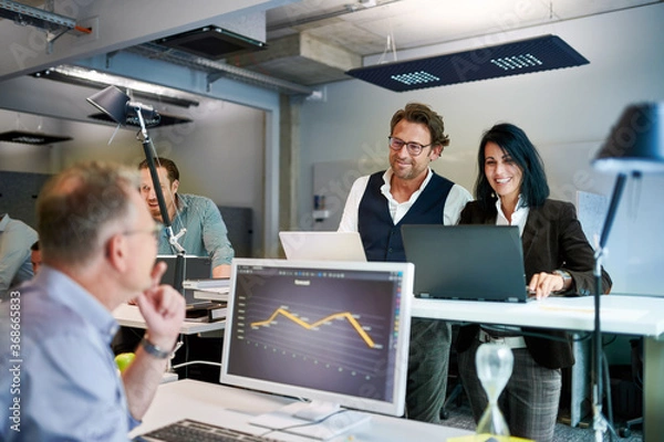 Obraz Smiling mature female and male colleagues discussing over graph while planning strategy at workplace
