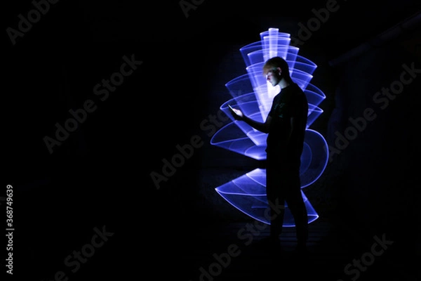 Fototapeta Silhouette of a man using a smartphone. Abstract shape at lightpainting with a light saber. Face illuminated by screen.	