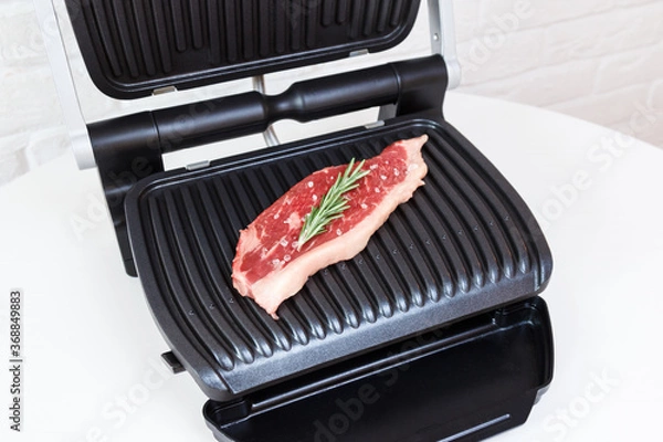 Fototapeta A piece of fresh meat with coarse salt and a sprig of rosemary. Juicy beef steak on the electric grill.