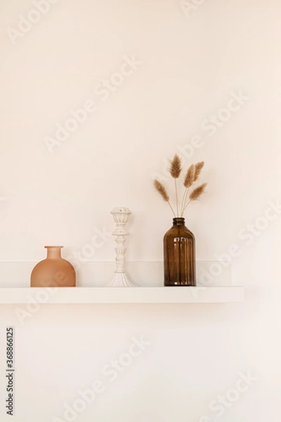 Fototapeta White shelf on white wall with bottle, vase, fluffy pom pom plants, candlestick. Minimal modern interior design concept.