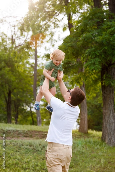 Obraz Young father and little son. Dad throws his son up against the background trees. Walk in park. Family life.