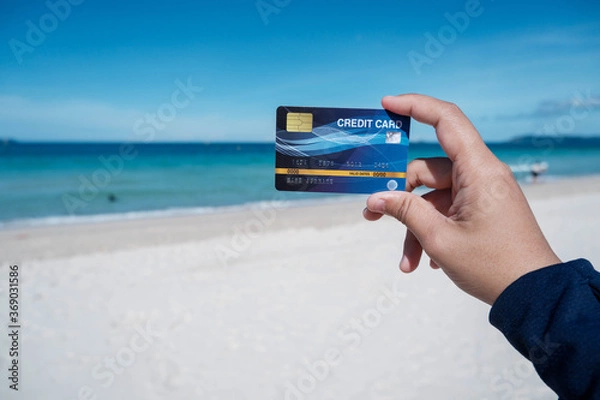 Fototapeta The hand holding a credit card that holds up in the sky, behind the sea is the concept of using a credit card for tourism and rest.