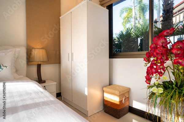 Fototapeta Interior design of house home, villa, condo, apartment feature white wardrobe, bedside table, lamp, basket, window glass and artificial flower in bedroom