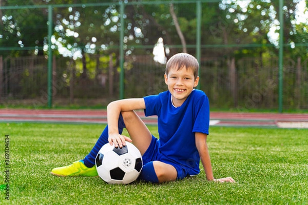 Fototapeta A little boy sits on a green soccer field, a soccer ball