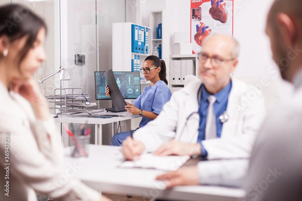 Fototapeta Medical nurse looking at x-ray. Senior doctor wearing white coat and stethoscope during consultation of couple in hospital office. Pair at medical check.