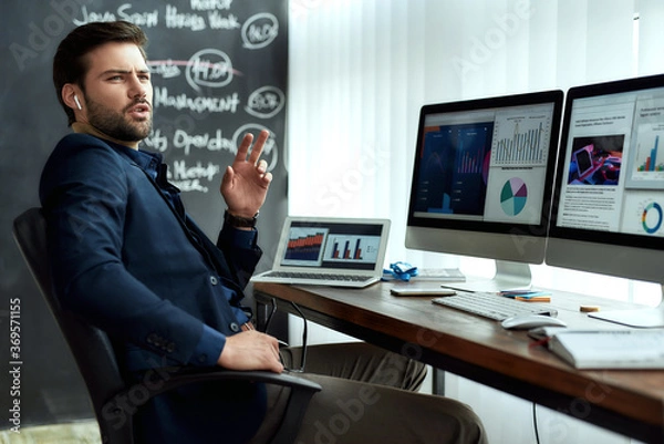 Fototapeta Young confident businessman in wireless earphones talking with client or partner, discussing sales report while working with statistical data in the modern office