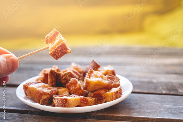 Fototapeta Hand holding a piece of fried pork rinds called “Torreznos” with a toothpick on a dark wooden table. Typicall spanish snacks to eat in a bar. Photo with selective focus. 