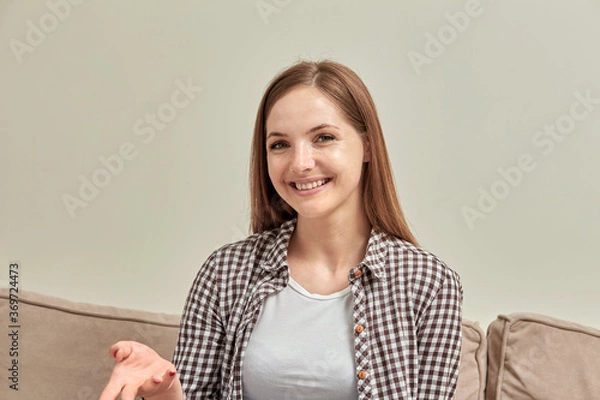 Fototapeta Woman portrait. Girl sitting on the couch looks at the camera and waves her hand