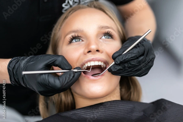Fototapeta Dentist examining patient teeth with a mouth mirror and dental excavator. Close-up view on the woman's face