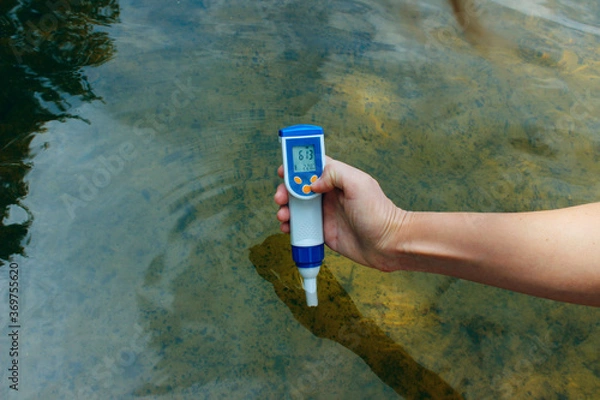 Fototapeta A woman's hand holds a waterproof tester pen and measures the total concentration of dissolved salts (TDS) and the temperature in the river. People, science concept. 